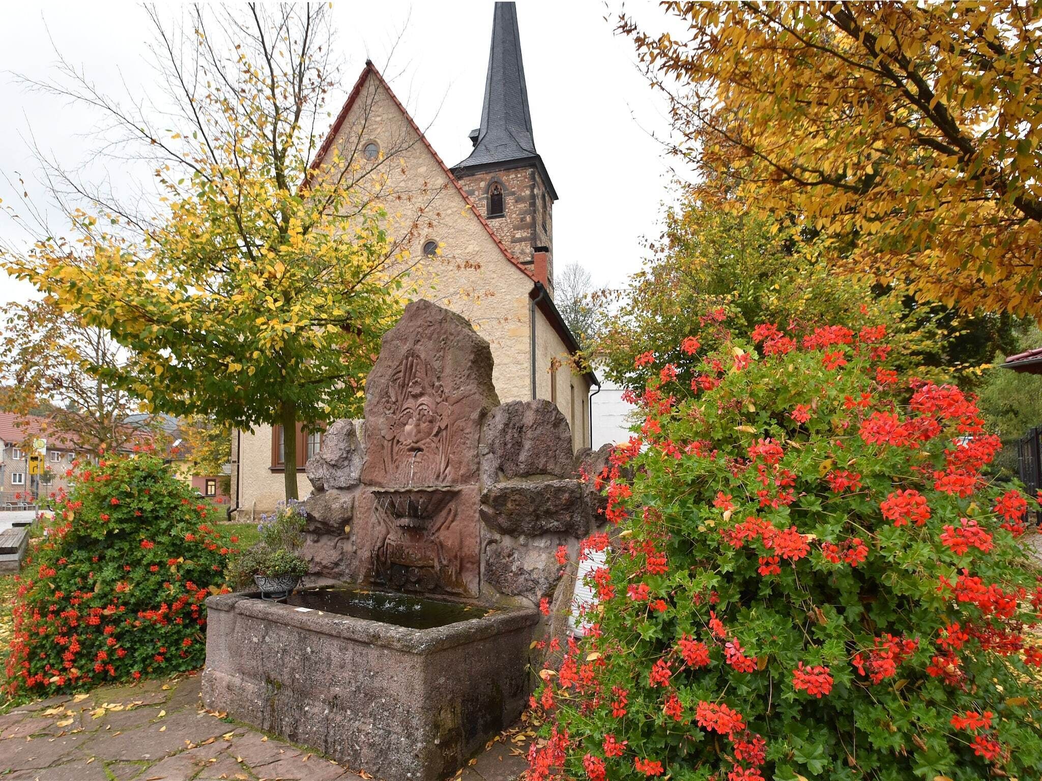 Eine Steinfontäne mit einer geschnitzten Figur, umgeben von roten Blumen und einer Kirche im Hintergrund.