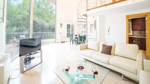 A spacious living room with a large TV, a glass coffee table, and a sofa. There's a staircase leading to an upper level with a balcony. - Villa in Alcúdia near Sandy Beach (Alcudia)