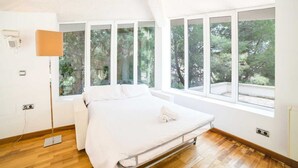 A modern bedroom with a large bed, multiple windows, and a wooden floor. - Villa in Alcúdia near Sandy Beach (Alcudia)