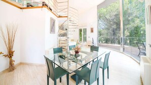 A modern dining area with a glass table and chairs, a spiral staircase, and large windows offering a view of trees. - Villa in Alcúdia near Sandy Beach (Alcudia)