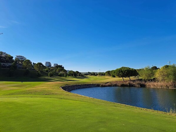 A golf course with a pond, green grass, and modern buildings in the background.