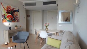 A modern living room with a sofa, a blue armchair, a small round table, and a dining area with a white table and chairs. - Golfside Luxury at Las Colinas (Orihuela Costa-Alicante)