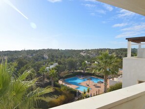 A pool area with multiple swimming pools, surrounded by palm trees and lounge chairs, with a view of a hilly landscape in the background.