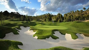 A golf course with sand bunkers, green fairways, and a forested hill in the background.