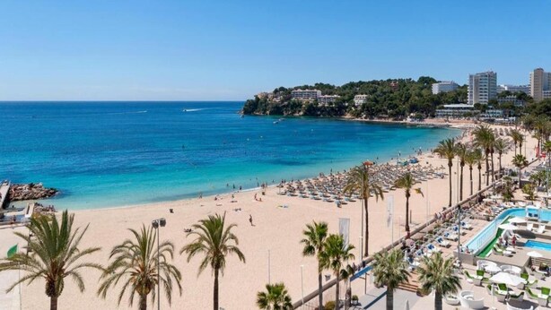Ein Strand mit klarem, blauem Wasser, Palmen und einer Stadtlandschaft im Hintergrund.