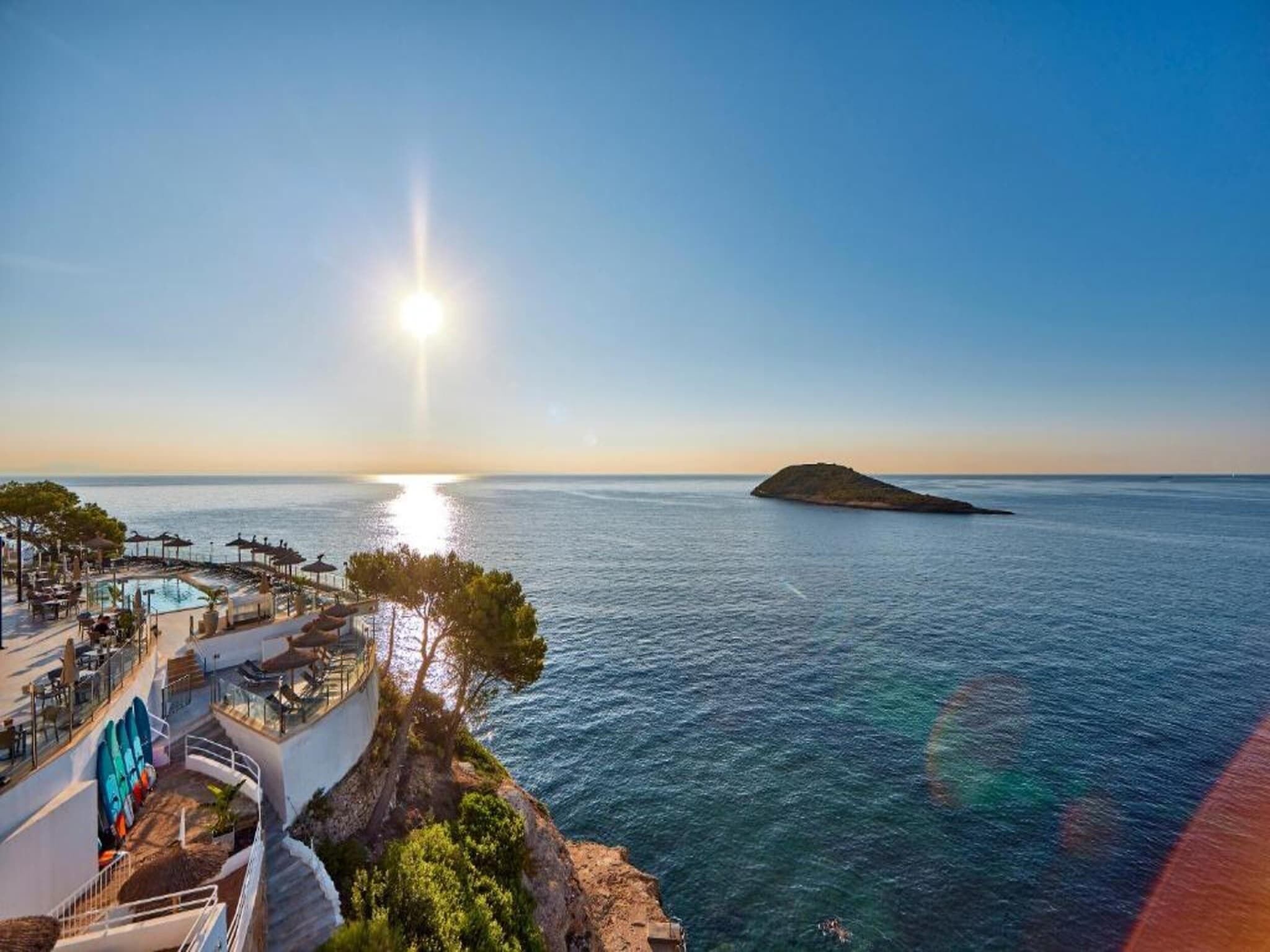 A coastal hotel with a pool area, outdoor seating, and a view of the sea and a small island.