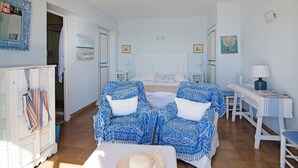 Una habitación con una cama, dos sillones azules estampados, una mesita, un armario blanco y un espejo con marco azul.