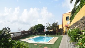 Una piscina rodeada de césped artificial, con un muro de piedra y un edificio amarillo al fondo.