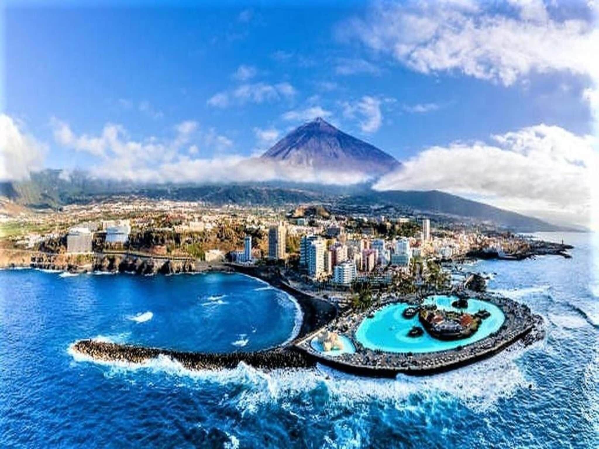 Un paisaje urbano costero con una montaña destacada al fondo, un puerto deportivo con barcos y una combinación de edificios residenciales y comerciales.