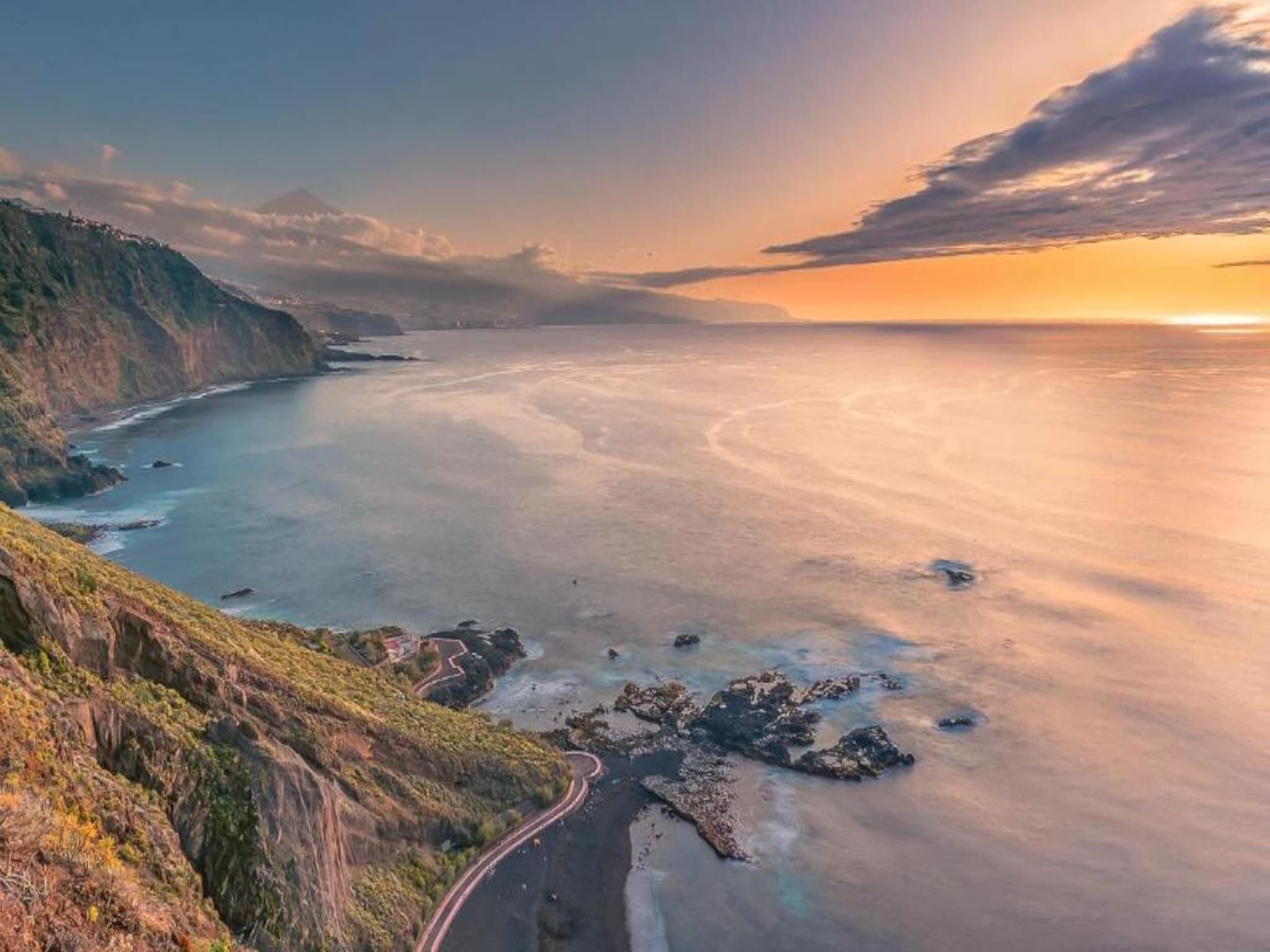Un paysage côtier avec une route sinueuse qui longe les falaises, une vue dégagée sur la mer et un coucher de soleil à l’horizon.