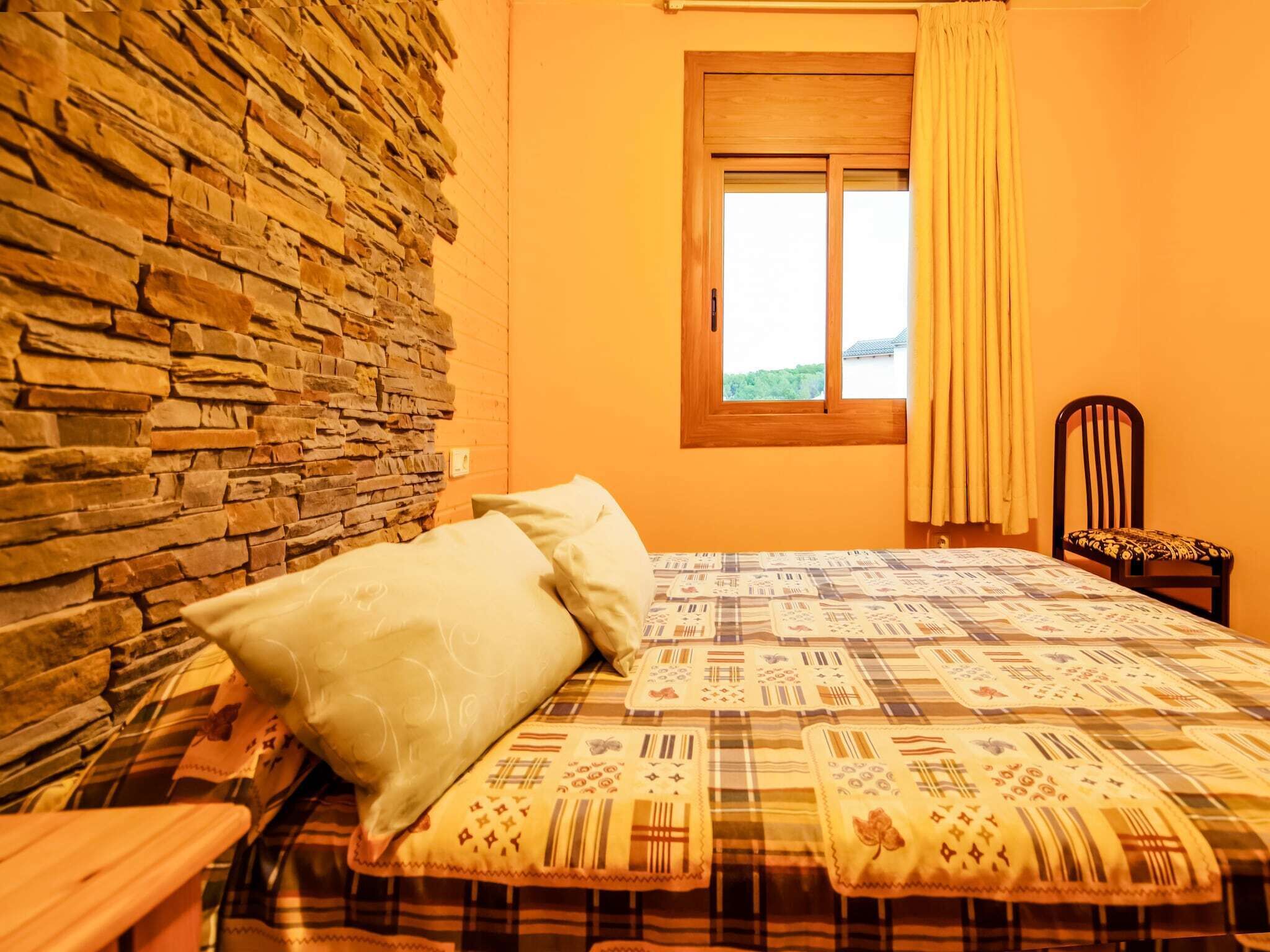 A room with a stone wall, a bed with patterned bedding, a window with curtains, and a chair.
