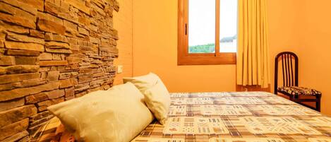 A room with a stone wall, a bed with patterned bedding, a window with curtains, and a chair.