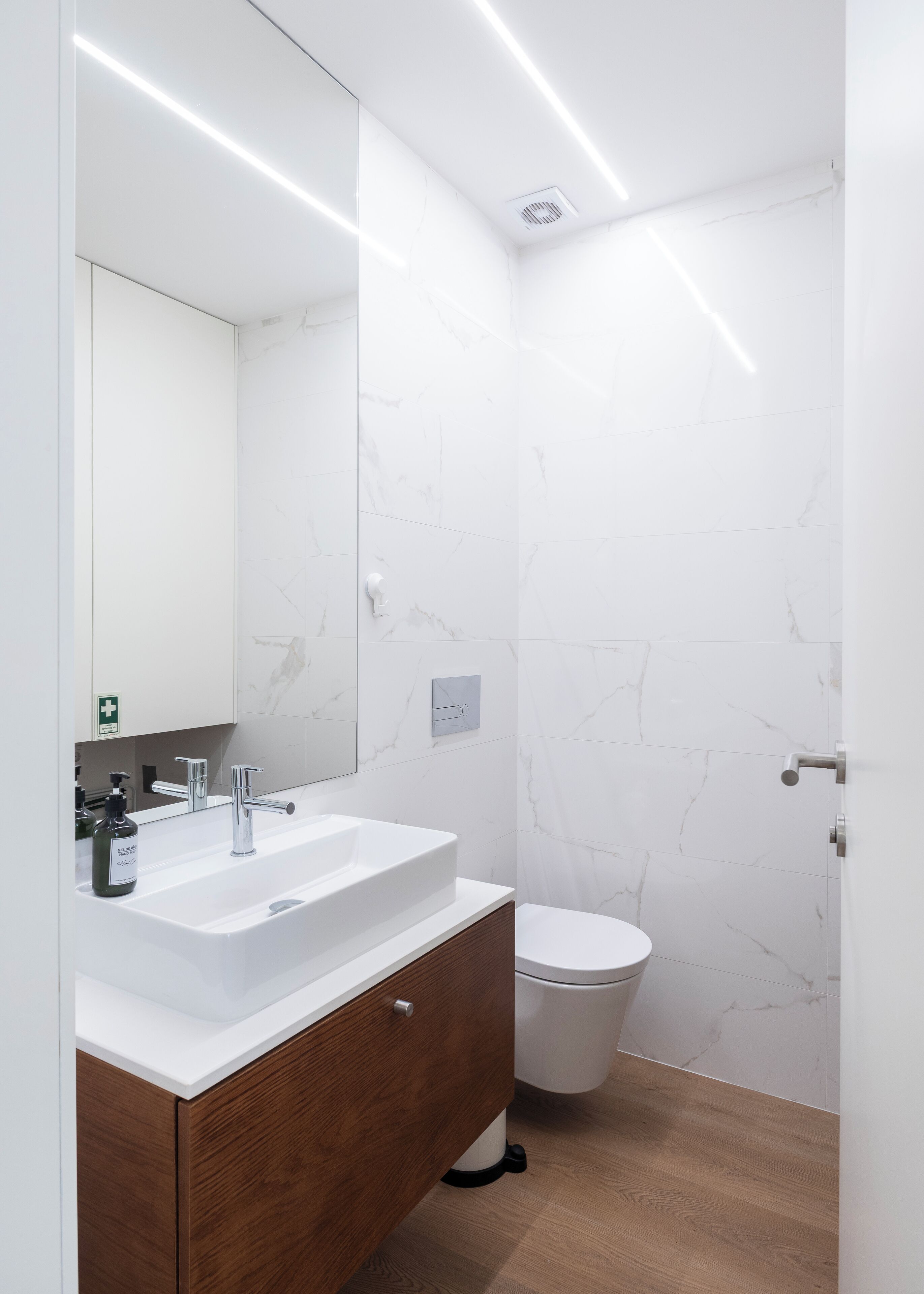 A modern bathroom with a white marble wall, a wooden vanity, a white sink, a mirror, and a toilet.