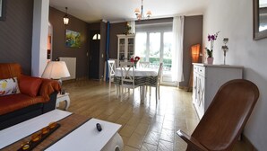 Una sala de estar con un sofá, una mesa de centro y una zona de comedor con una mesa y sillas. Se aprecia un aparador con objetos decorativos y vistas al exterior.