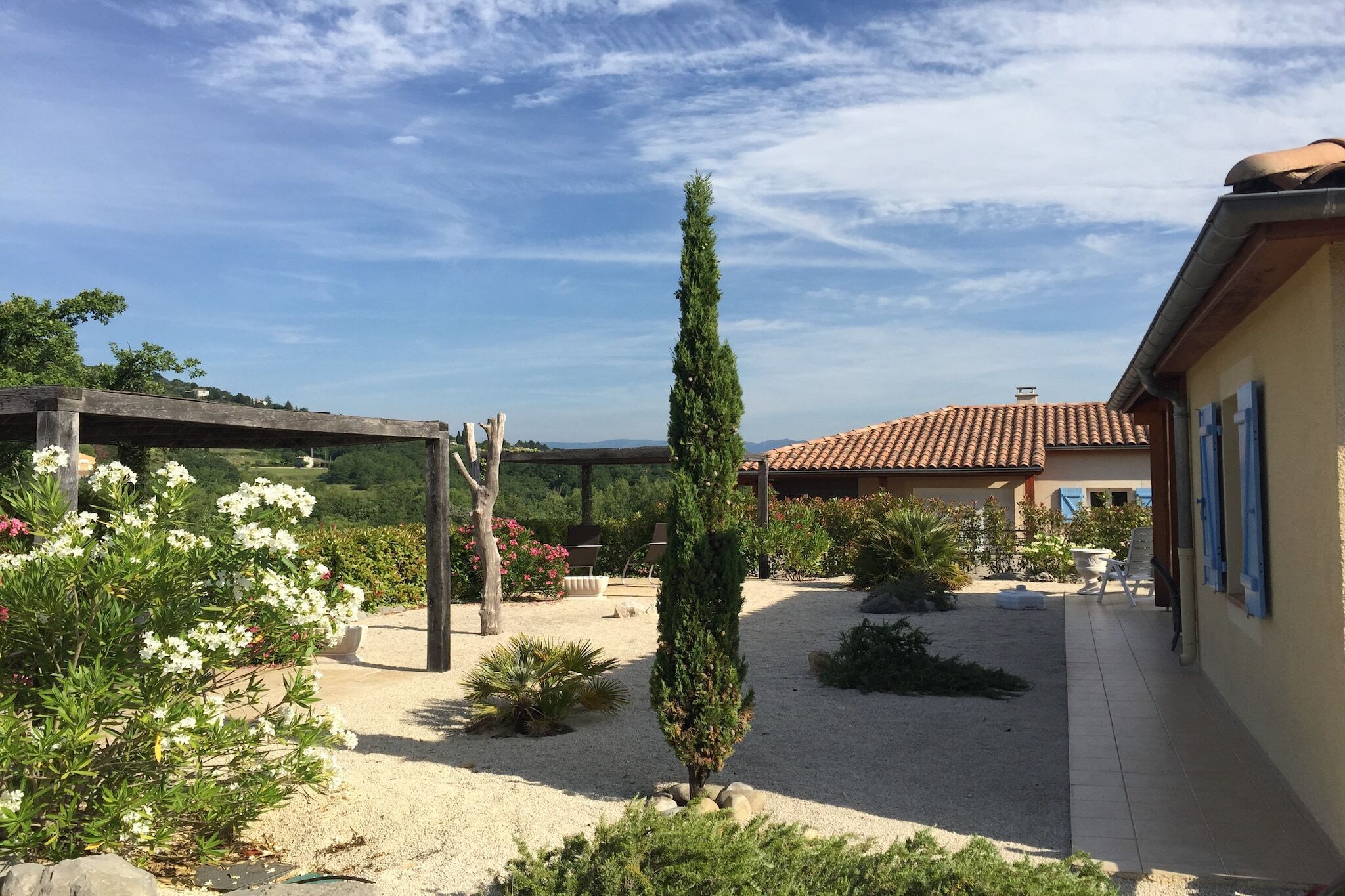 A garden with a pergola, a tall coniferous tree, and a building with a tiled roof.