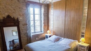 A bedroom with a stone wall, wooden paneling, a large bed with white linens, two bedside tables with lamps, and a window with curtains. - Holiday Home in Béziers with Pool and Vineyard (Murviel les Béziers)