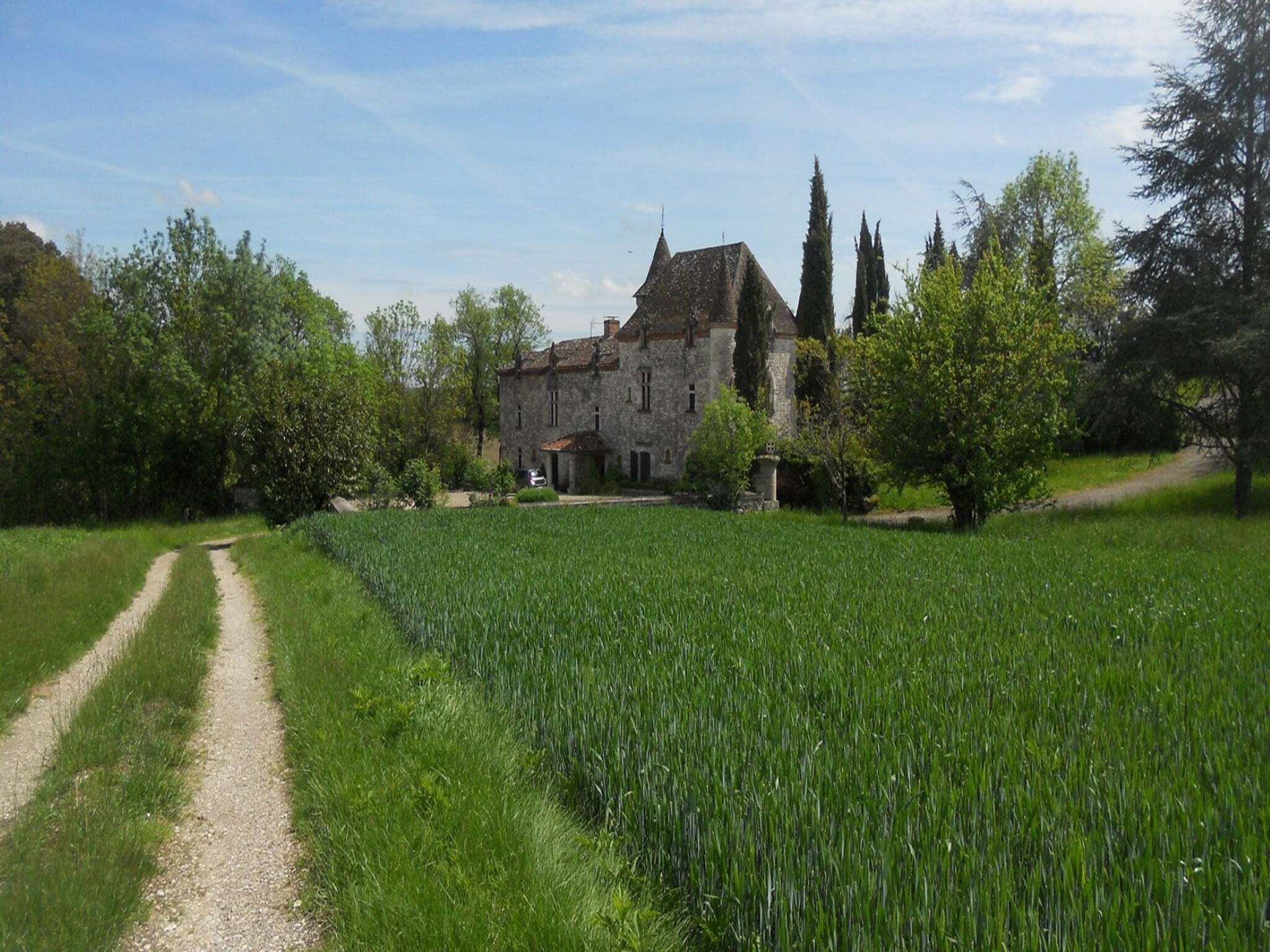 Un grand bâtiment en pierre, doté d’une façade symétrique, entouré d’un champ verdoyant et luxuriant, et auquel mène un chemin de gravier.