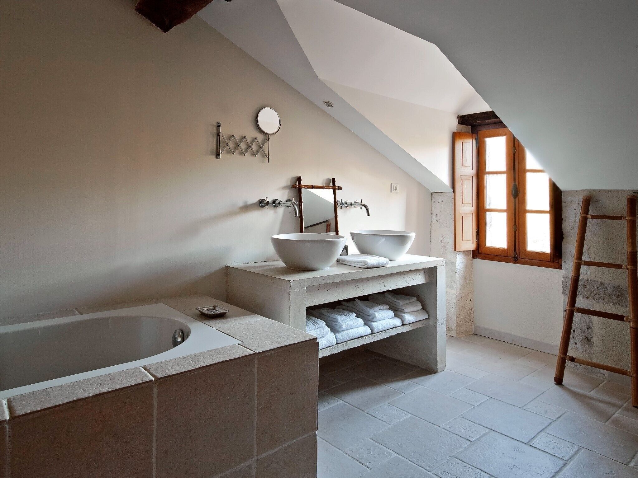 Een moderne badkamer met een vrijstaand bad, twee grote witte waskommen, een spiegel en een raam met natuurlijk licht.