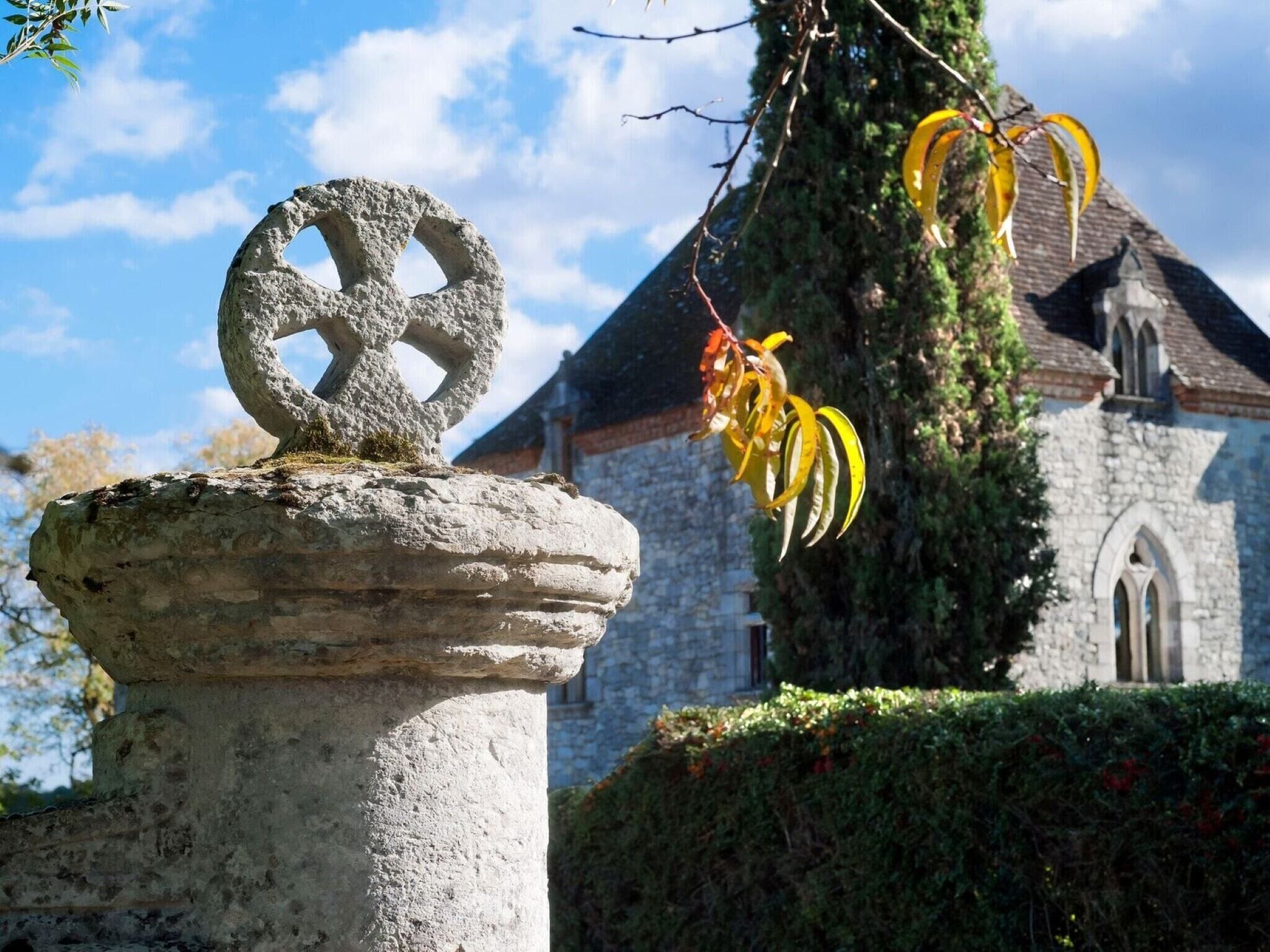 A stone monument with a circular design, a church with a tiled roof, and a tree with yellow leaves in the background.