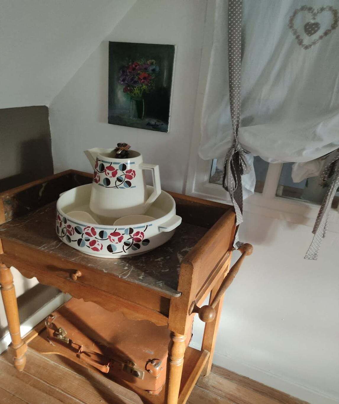 Une table en bois, sur laquelle sont posés un pichet et une théière blancs ornés de motifs floraux, un tableau représentant des fleurs accroché au mur et un ornement en forme de cœur sur le rideau.