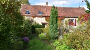 A traditional house with a red-tiled roof, a stone wall, and a garden with various plants and a tall tree.