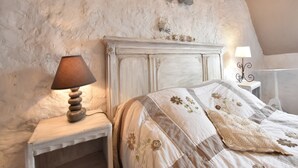 A bedroom with a rustic stone wall, a wooden nightstand with a lamp, a bed with floral bedding, and a decorative bow on the wall.