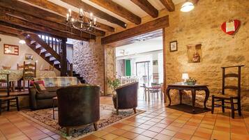A rustic interior with exposed wooden beams, a chandelier, a stone wall, and a staircase leading to another room.