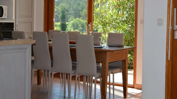 A dining area with a wooden table and chairs, a kitchen with cabinets, and a large window with a view of greenery. - Villa Overlooking Les Vans Centre (Les Vans)