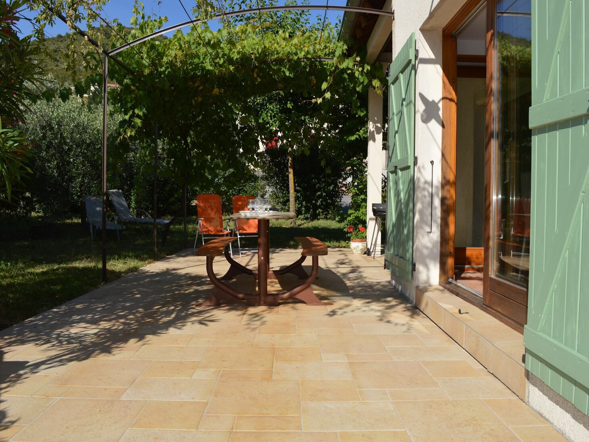 An outdoor patio with a table and chairs, surrounded by greenery and a building with a green door.