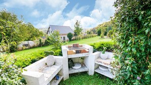 Een betonnen terras buiten met een barbecue, omgeven door weelderige begroeiing en een huis op de achtergrond.