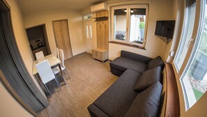 Un salon moderne avec un canapé d’angle gris, une table à manger avec des chaises, un coin cuisine et une grande fenêtre qui laisse entrer la lumière naturelle.