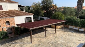 A patio with a metal table and stone flooring, surrounded by greenery and a white building with a tiled roof. - Villa Pinna (San Teodoro)