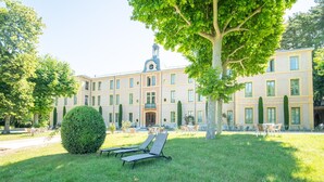 Un bâtiment à la façade symétrique, doté de nombreuses fenêtres et d’une entrée centrale. On y trouve des espaces extérieurs aménagés avec des tables et des chaises, ainsi qu’un grand arbre qui offre de l’ombre.