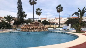 A swimming pool with a central rock feature, surrounded by palm trees and a red pathway. - Charming 3-bedroom townhome with AC, WiFi in lovely Retamar (Retamar)