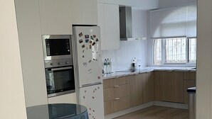 A modern kitchen with white cabinetry, a built-in oven, and a refrigerator. There is a dining table with a blue tablecloth and a chair. The floor is wooden, and there is a window providing natural light. - Charming 3-bedroom townhome with AC, WiFi in lovely Retamar (Retamar)
