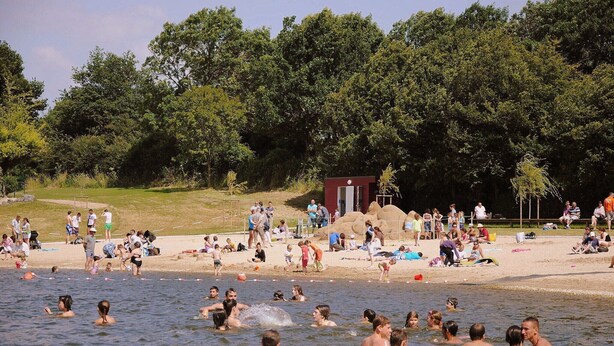 Une plage bondée où l’on voit des gens nager et bronzer, une petite construction et, au loin, une colline boisée.