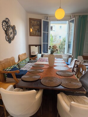 A dining area with a long table, chairs, and a view through open windows.