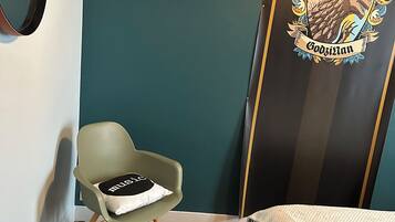 A room with a bed, a green chair, a wooden floor, a round stool, and a door with a coat of arms.