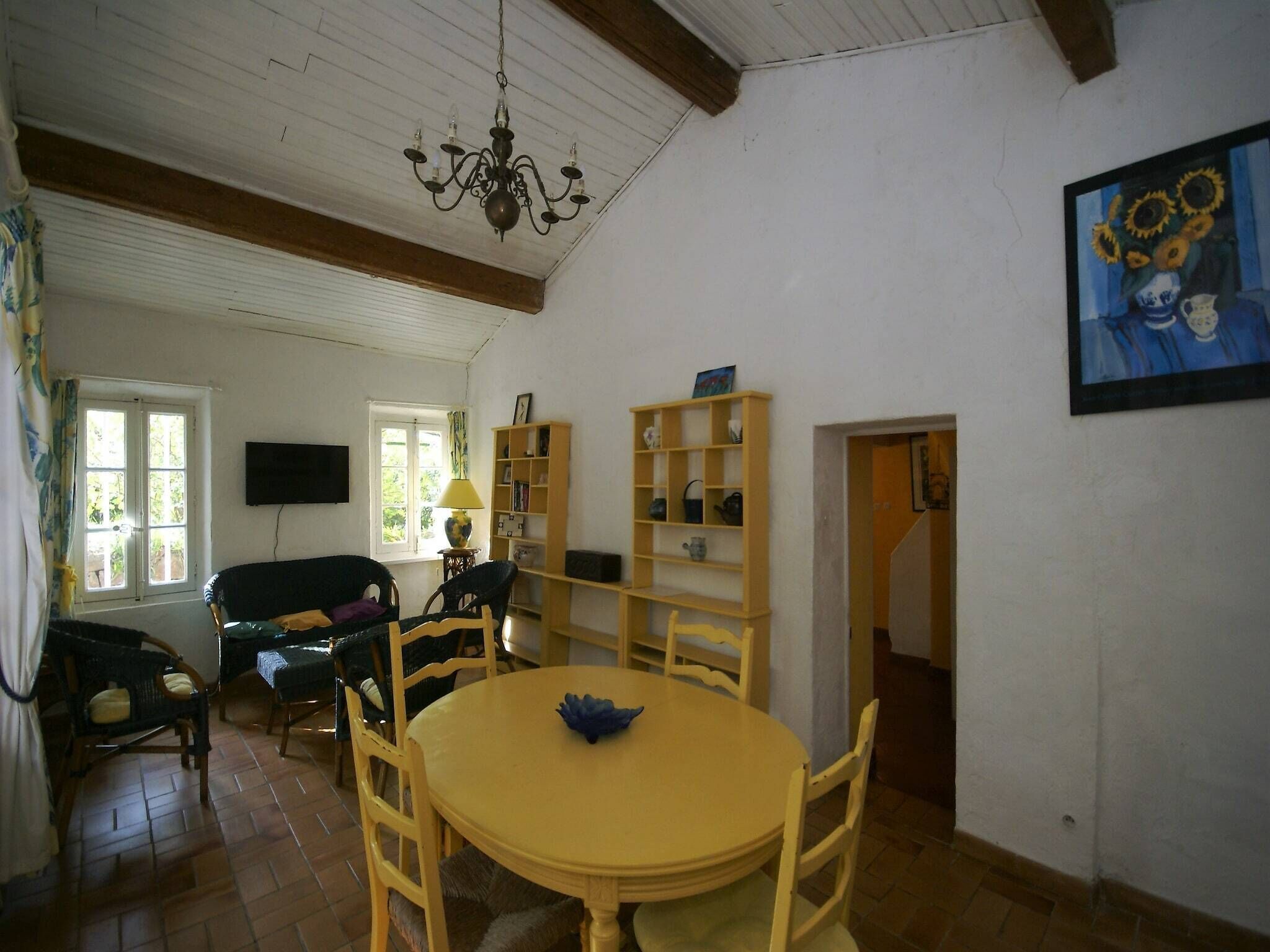 A dining area with a round yellow table, chairs, a chandelier, a bookshelf, and a painting on the wall.