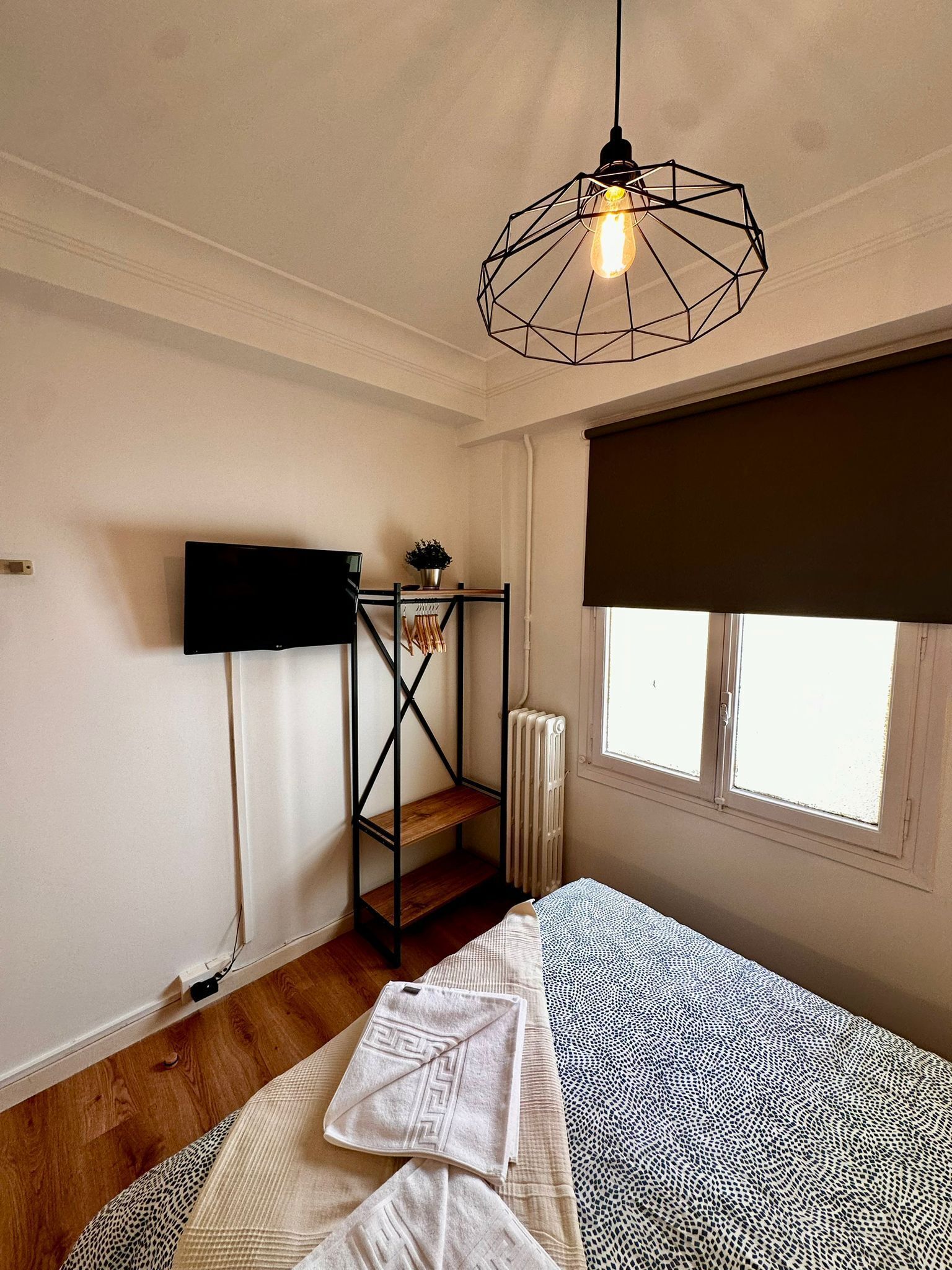 Un dormitorio con cama, lámpara colgante, radiador y ventana con persiana.