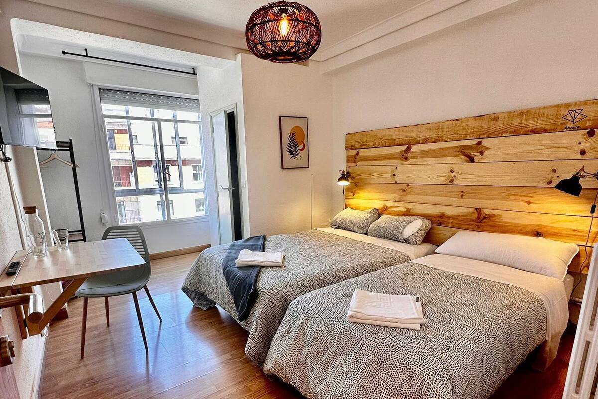 A bedroom with a wooden headboard, two beds with patterned bedding, a desk with a chair, and a hanging light fixture.