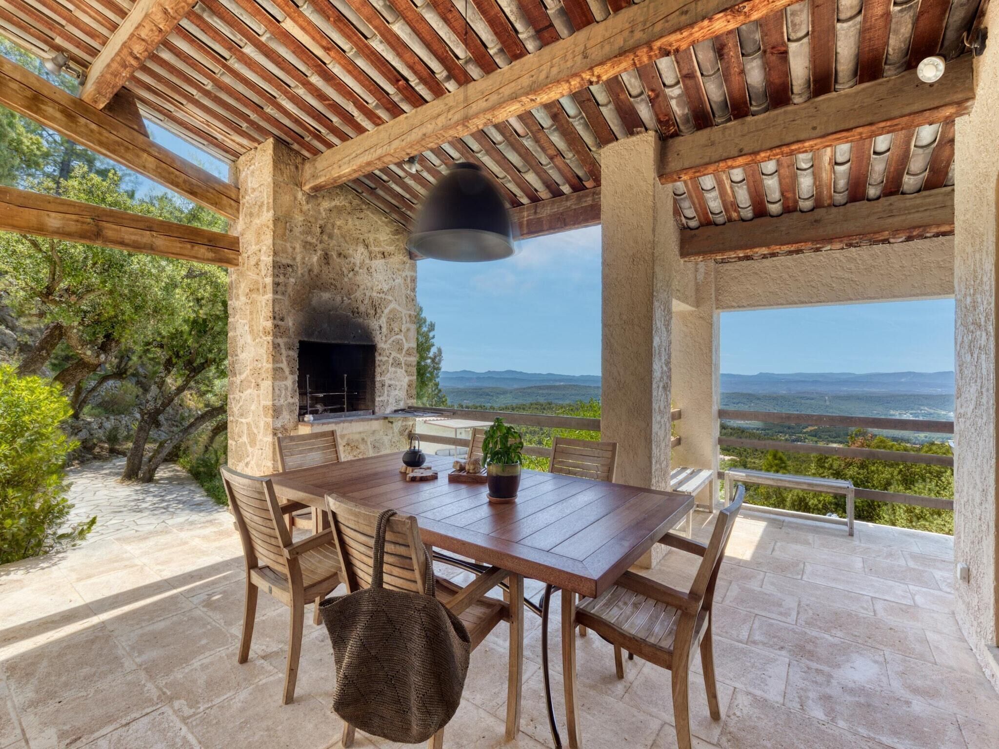 An outdoor patio with a stone fireplace, wooden table, chairs, and a view of the landscape.
