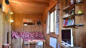 Ein gemütliches Blockhaushaus-Interieur mit Holzboden, einer gemusterten Couch, einem kleinen Tisch, einem Fernseher und einem Bücherregal.