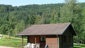 A wooden cabin with a tiled roof, a porch with white plastic chairs, and a view of a forested hillside. - Ventron Chalet with Hill Views (Ventron)
