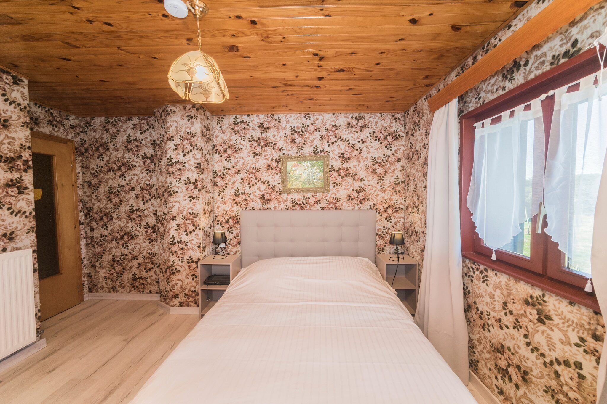 Un dormitorio con papel tapiz floral, techo de madera, cama con colcha blanca, dos mesitas de noche con lámparas y una ventana con cortinas blancas.