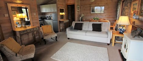 Un acogedor interior de cabaña de madera con un sofá blanco, una silla de madera, una mesita y una zona de cocina al fondo.