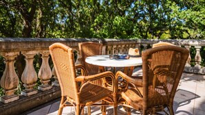 Des chaises et une table en osier sur une terrasse extérieure, avec une balustrade en pierre et des arbres en arrière-plan.