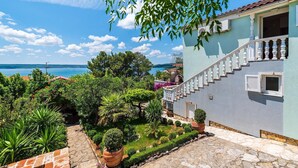 Une propriété située sur la côte, dotée d’un chemin en pierre, d’un jardin luxuriant et d’un escalier menant à un bâtiment bleu clair avec un balcon.