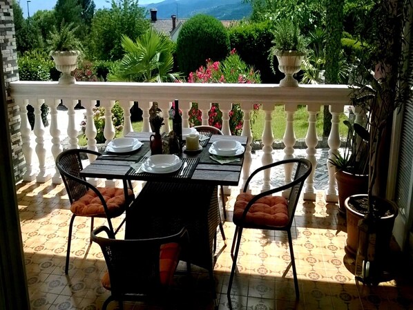 A balcony with a table set for dining, surrounded by chairs with cushions, and potted plants.