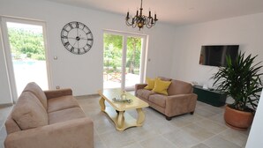 A living room with beige sofas, a coffee table, a wall clock, and a TV. - Villa in Tijarica with Private Pool (Tijarica)